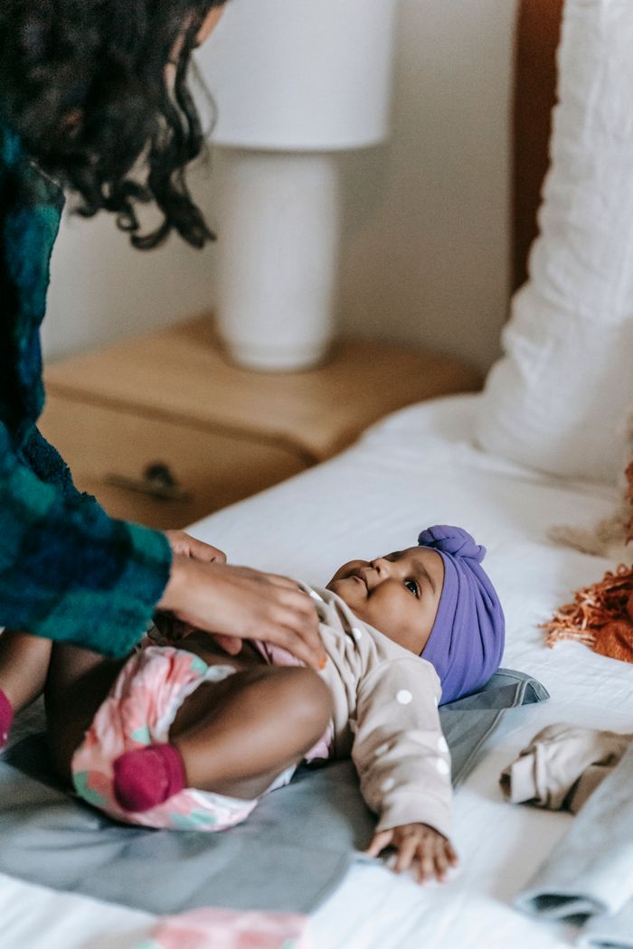 portfolio-04 Unrecognizable African American mother putting clothes on cute black baby with headband lying on soft bed in bedroom at home